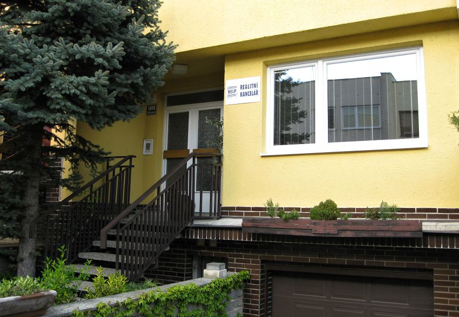 Žlutý dům s cedulí HELP realitní kancelář, vstup po schodech vedle smrku.
